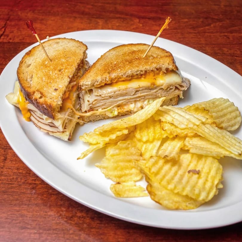 A grilled turkey and cheese sandwich, cut in half, with a side of ridged potato chips on a white plate.