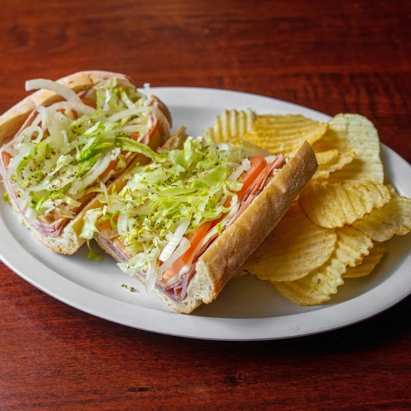 Two halves of a Frankie's Pizza II sub sandwich with lettuce, tomato, and onions, served with potato chips.